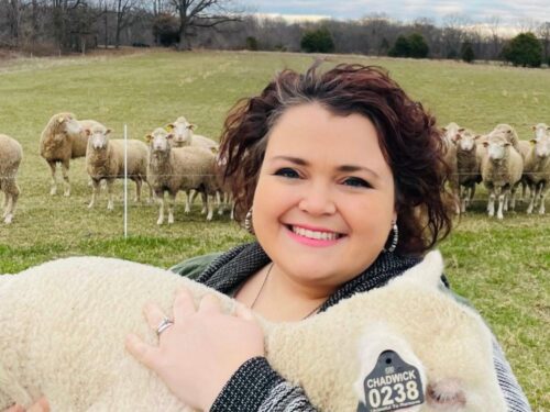 Kelsey Chadwick, author of Where Else Could I go holds a sheep with field and sheep grazing in background